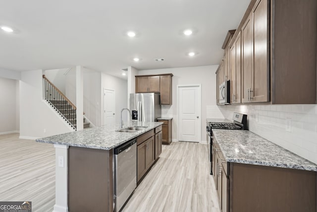 kitchen with sink, stainless steel appliances, light stone counters, an island with sink, and light wood-type flooring