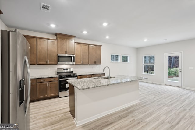 kitchen with light wood-type flooring, light stone counters, stainless steel appliances, sink, and an island with sink