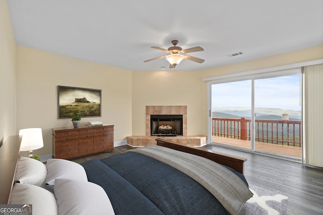 bedroom featuring a tiled fireplace, access to outside, dark wood-type flooring, a mountain view, and ceiling fan