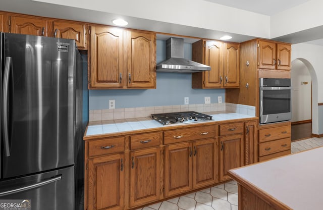 4909 Perry Rd, Claxton, GA 30417 - kitchen with appliances with stainless steel finishes, wall chimney exhaust hood, brown cabinetry, arched walkways, and tile countertops