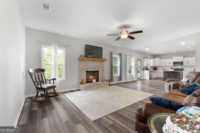 living room featuring dark hardwood / wood-style flooring, a wealth of natural light, ceiling fan, and a stone fireplace