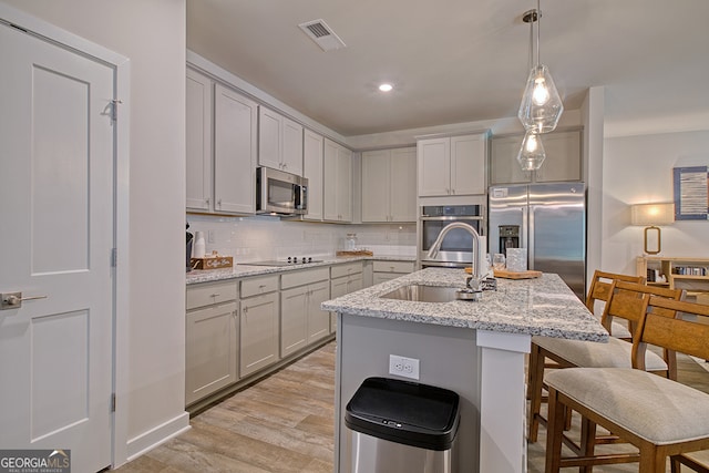 kitchen featuring light stone counters, an island with sink, sink, stainless steel appliances, and light wood-type flooring