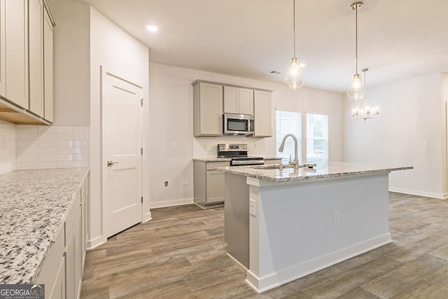 kitchen with a center island with sink, appliances with stainless steel finishes, light stone counters, light wood-type flooring, and a sink