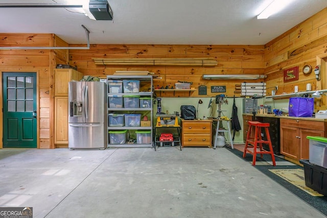 garage with a garage door opener, a workshop area, stainless steel refrigerator with ice dispenser, and wooden walls