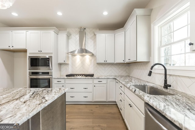 kitchen with white cabinets, wall chimney exhaust hood, stainless steel appliances, light wood-style floors, and a sink