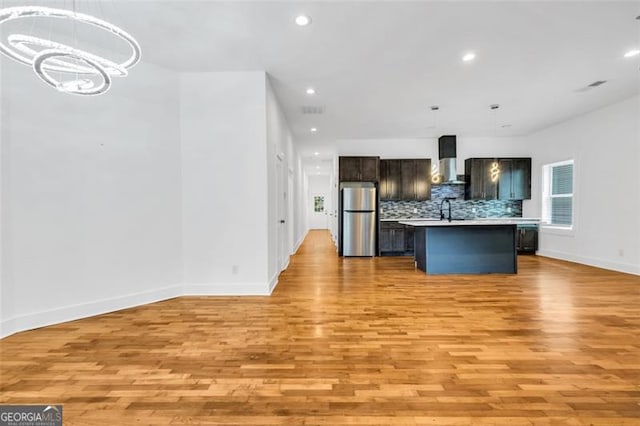 kitchen featuring stainless steel refrigerator, light wood-type flooring, decorative light fixtures, a center island with sink, and wall chimney range hood