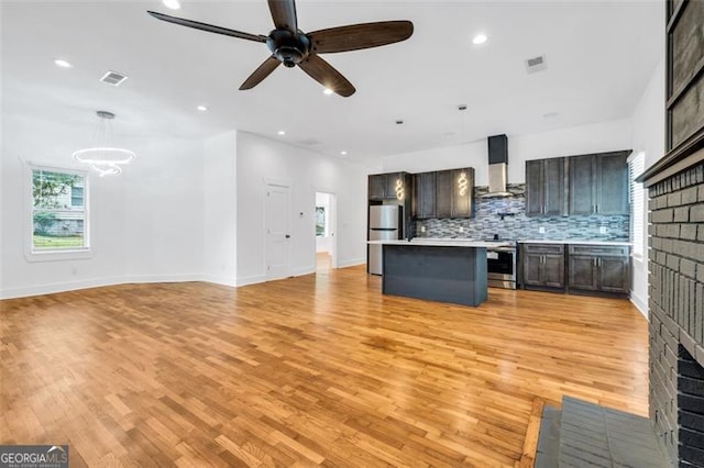 kitchen featuring hanging light fixtures, a kitchen island, wall chimney range hood, appliances with stainless steel finishes, and ceiling fan with notable chandelier