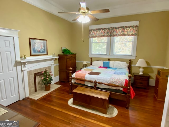 bedroom with ornamental molding, a tiled fireplace, ceiling fan, and dark wood-type flooring