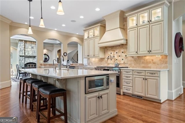 kitchen with premium range hood, a kitchen island with sink, cream cabinetry, stainless steel appliances, and light wood-type flooring