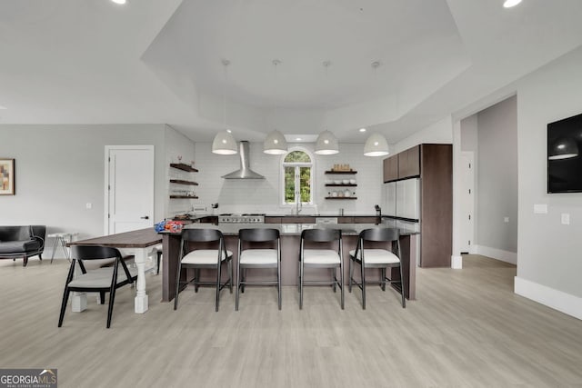 kitchen featuring a kitchen breakfast bar, wall chimney range hood, hanging light fixtures, light hardwood / wood-style floors, and dark brown cabinetry