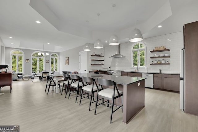 kitchen with appliances with stainless steel finishes, backsplash, wall chimney exhaust hood, decorative light fixtures, and light hardwood / wood-style flooring