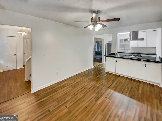 kitchen featuring white cabinets, wall chimney exhaust hood, hardwood / wood-style flooring, appliances with stainless steel finishes, and ceiling fan with notable chandelier