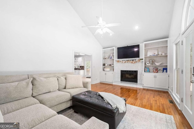 living room with light wood-type flooring, a fireplace, ceiling fan, high vaulted ceiling, and built in shelves