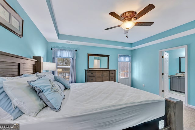 carpeted bedroom featuring connected bathroom, a tray ceiling, ceiling fan, and multiple windows