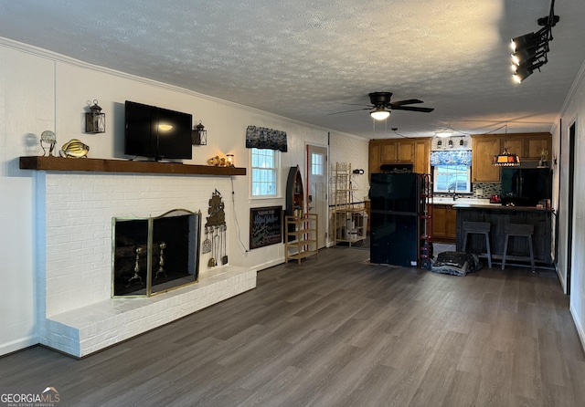 living room with a brick fireplace, ceiling fan, dark wood-type flooring, a textured ceiling, and ornamental molding