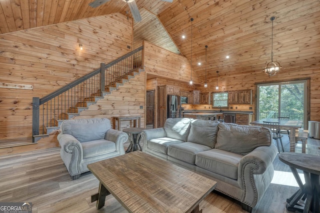 living room featuring high vaulted ceiling, wooden walls, light hardwood / wood-style flooring, and ceiling fan with notable chandelier