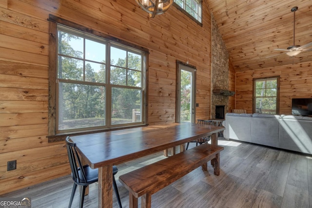 dining room featuring dark hardwood / wood-style floors, ceiling fan with notable chandelier, wood walls, high vaulted ceiling, and a stone fireplace