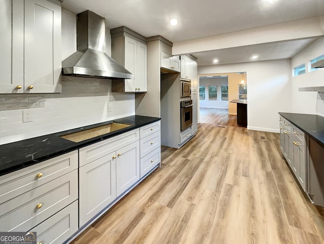 kitchen with appliances with stainless steel finishes, gray cabinetry, light hardwood / wood-style floors, wall chimney exhaust hood, and dark stone counters