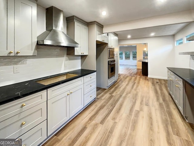 kitchen with light wood-type flooring, dark countertops, stainless steel appliances, and wall chimney exhaust hood