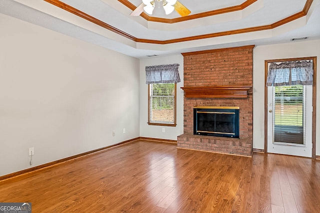 unfurnished living room featuring a tray ceiling, hardwood / wood-style flooring, a brick fireplace, crown molding, and ceiling fan