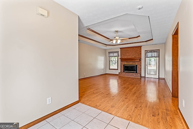 unfurnished living room featuring a brick fireplace, a textured ceiling, a tray ceiling, ceiling fan, and light hardwood / wood-style flooring