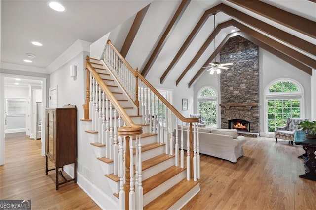 stairs featuring ceiling fan, beam ceiling, wood-type flooring, a stone fireplace, and high vaulted ceiling