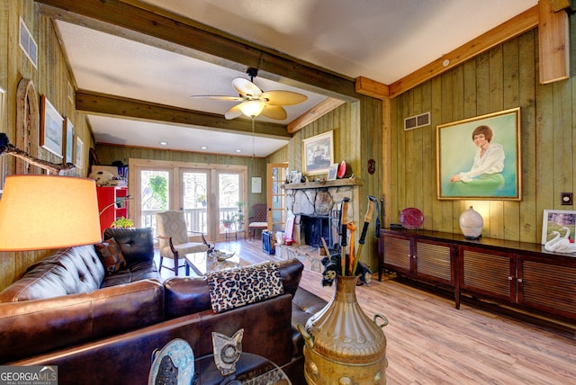 living room with ceiling fan, beam ceiling, a stone fireplace, wooden walls, and light hardwood / wood-style flooring