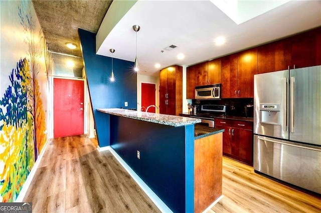 kitchen featuring appliances with stainless steel finishes, an island with sink, tasteful backsplash, light hardwood / wood-style flooring, and decorative light fixtures