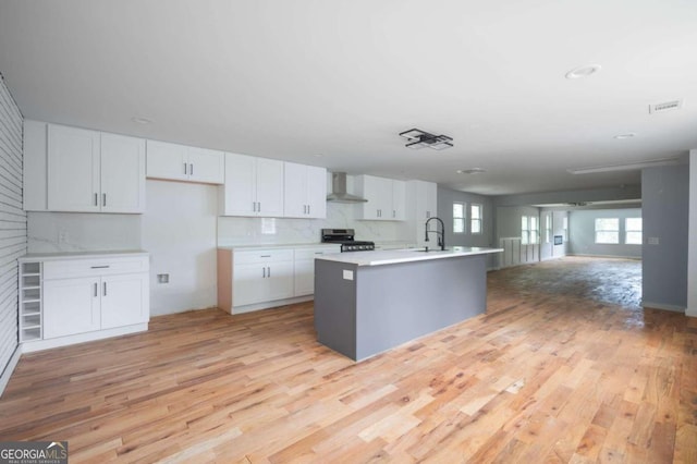 kitchen with white cabinetry, a kitchen island with sink, stainless steel gas range oven, and wall chimney range hood