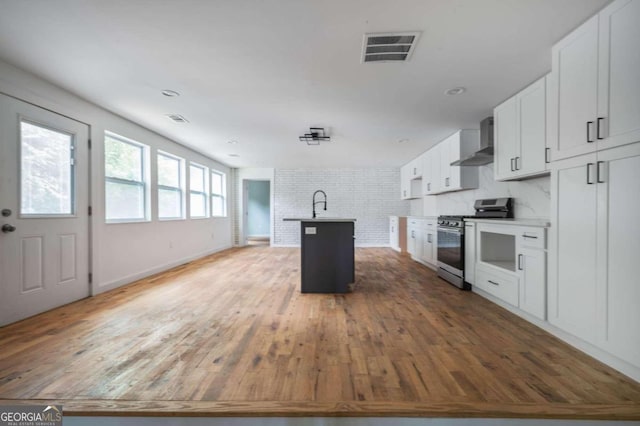 kitchen featuring wall chimney range hood, brick wall, a center island with sink, white cabinets, and stainless steel range with gas stovetop