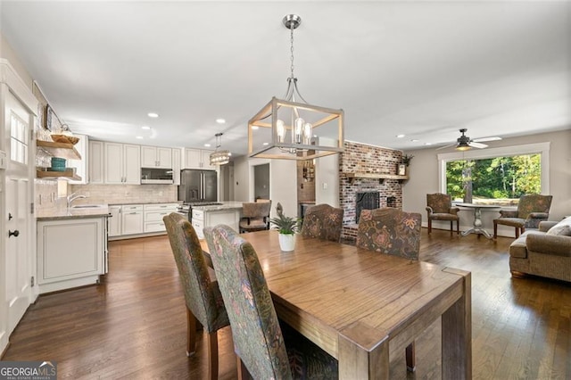 dining area featuring ceiling fan with notable chandelier, sink, a brick fireplace, and dark hardwood / wood-style floors