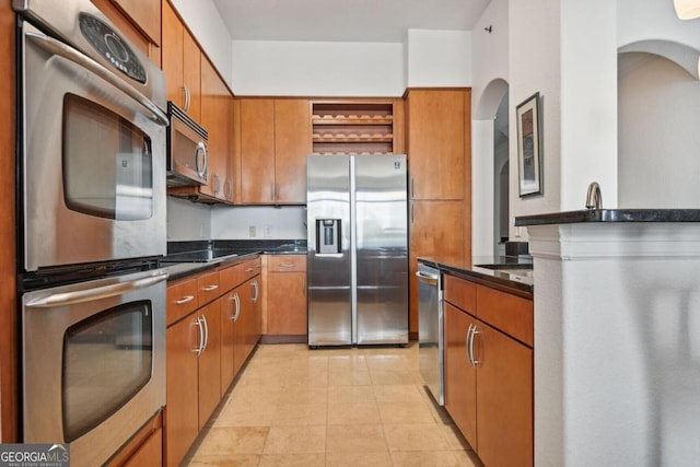 kitchen with brown cabinets, light tile patterned floors, open shelves, appliances with stainless steel finishes, and dark stone counters