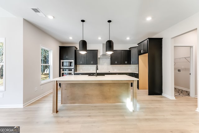 kitchen featuring appliances with stainless steel finishes, wall chimney range hood, sink, a center island with sink, and light hardwood / wood-style floors