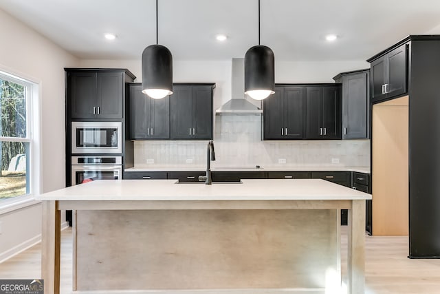 kitchen with wall chimney range hood, decorative backsplash, pendant lighting, a kitchen island with sink, and appliances with stainless steel finishes