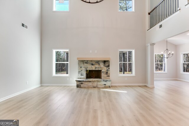 unfurnished living room featuring light wood-type flooring, a towering ceiling, ornate columns, a notable chandelier, and a stone fireplace