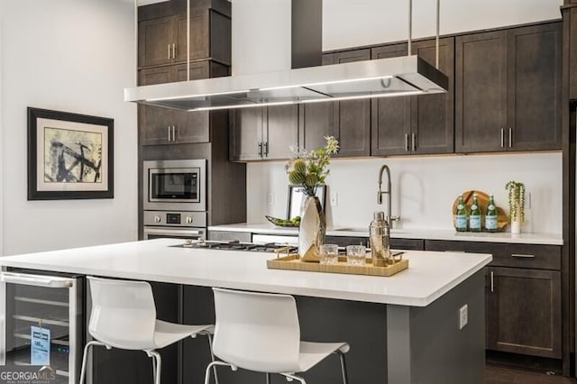 kitchen with a center island with sink, wine cooler, wall chimney exhaust hood, appliances with stainless steel finishes, and dark brown cabinets