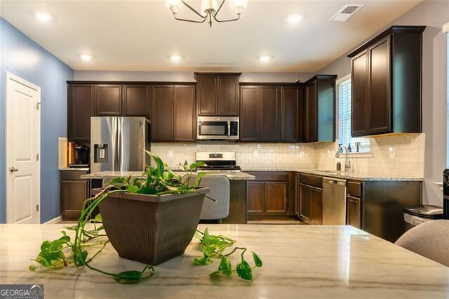 kitchen with light stone counters, a kitchen island, backsplash, appliances with stainless steel finishes, and an inviting chandelier