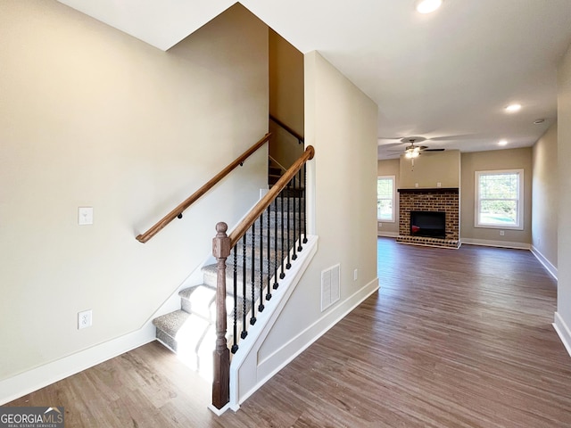 staircase with visible vents, a ceiling fan, a brick fireplace, wood finished floors, and baseboards