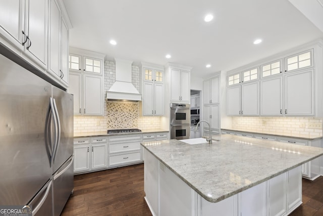 kitchen with stainless steel appliances, white cabinets, sink, an island with sink, and premium range hood