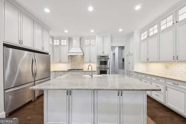 kitchen featuring stainless steel appliances, light stone counters, white cabinets, custom exhaust hood, and an island with sink