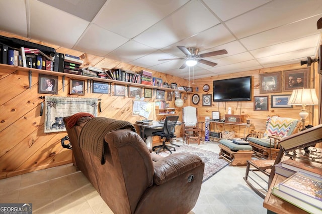 living room with ceiling fan, a paneled ceiling, and wood walls