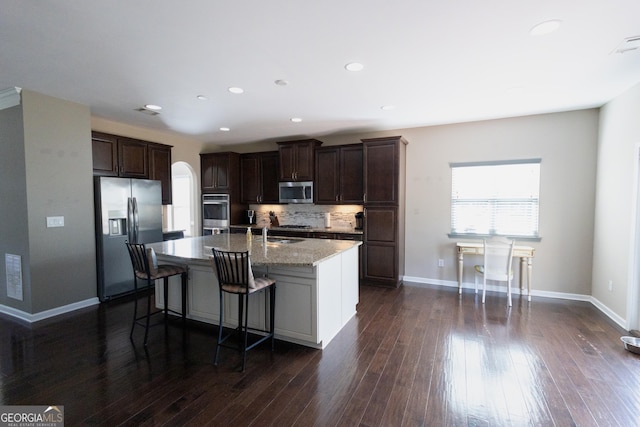 kitchen with dark wood-type flooring, stainless steel appliances, a center island with sink, backsplash, and a kitchen breakfast bar