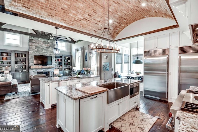 kitchen featuring light stone countertops, pendant lighting, a center island with sink, built in appliances, and ceiling fan with notable chandelier