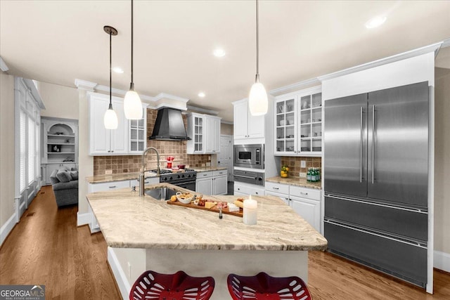 kitchen with built in appliances, wall chimney exhaust hood, an island with sink, and decorative light fixtures