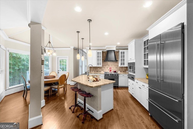 kitchen featuring wall chimney exhaust hood, light stone counters, a kitchen bar, a kitchen island with sink, and high end appliances