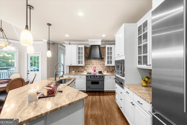 kitchen featuring wall chimney exhaust hood, sink, built in appliances, decorative light fixtures, and white cabinets