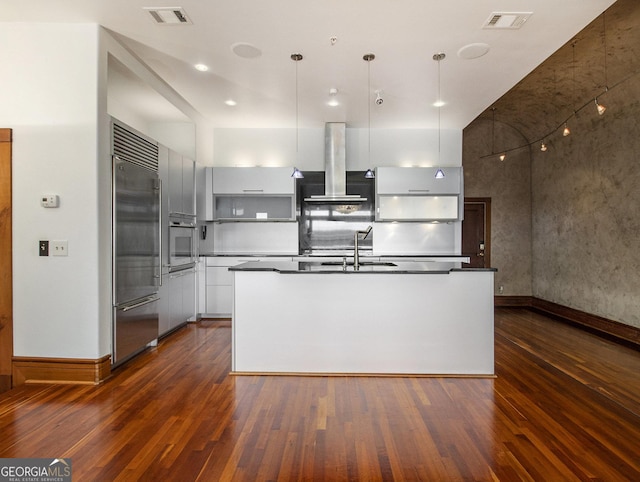 kitchen featuring a kitchen island with sink, white cabinets, built in refrigerator, wall chimney exhaust hood, and decorative light fixtures