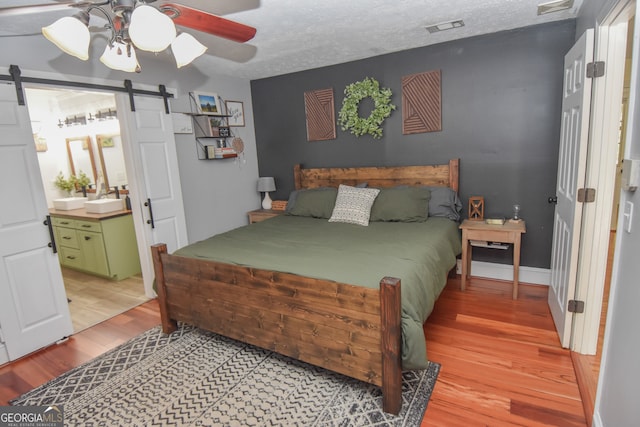 bedroom featuring ceiling fan, a textured ceiling, a barn door, light wood-type flooring, and ensuite bathroom