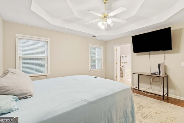 bedroom with ceiling fan, ensuite bath, wood-type flooring, and a tray ceiling