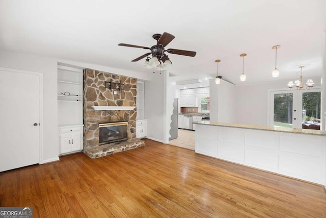 unfurnished living room with ceiling fan with notable chandelier, built in shelves, light hardwood / wood-style floors, and a stone fireplace